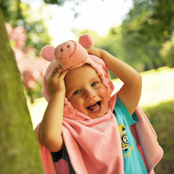 A young child smiling widely whilst wearing a light pink animal cape and touching the padded and embroidered pig nose