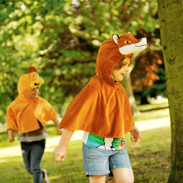 Child running around a park wearing an orange fox hooded cape with a child running in a chicken cape in the background. The side view of the fox costume shows the detailed embroidered fox facial features and fox ears